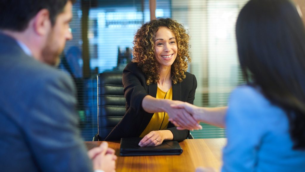 Una mujer de pelo rizado está sentada frente a un escritorio, sonríe y estrecha la mano de otra persona mientras un hombre la observa.