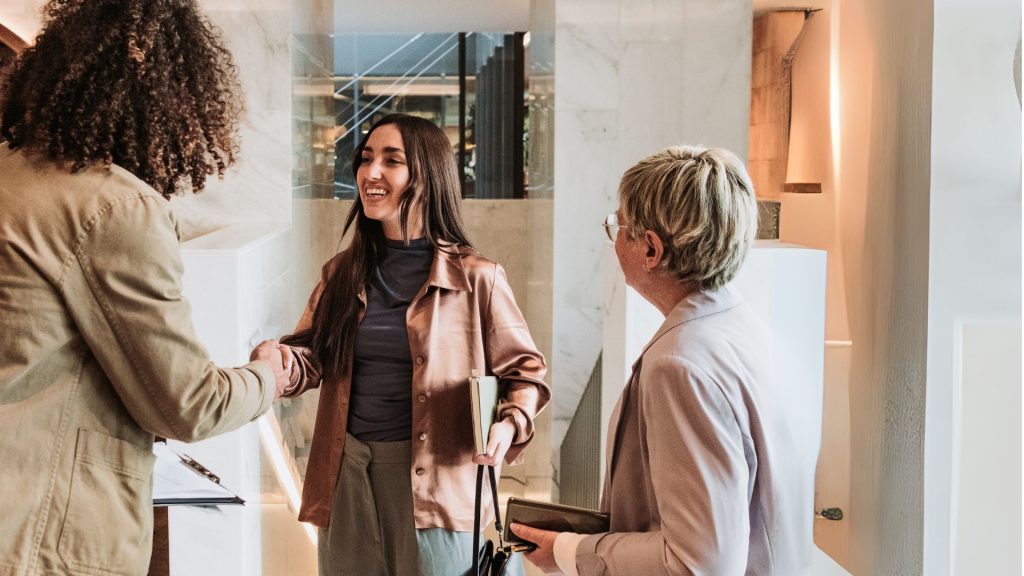 Una mujer sonríe y estrecha la mano de otra mujer, mientras otra mujer se encuentra cerca sosteniendo una cartera. Están en un interior luminoso y moderno