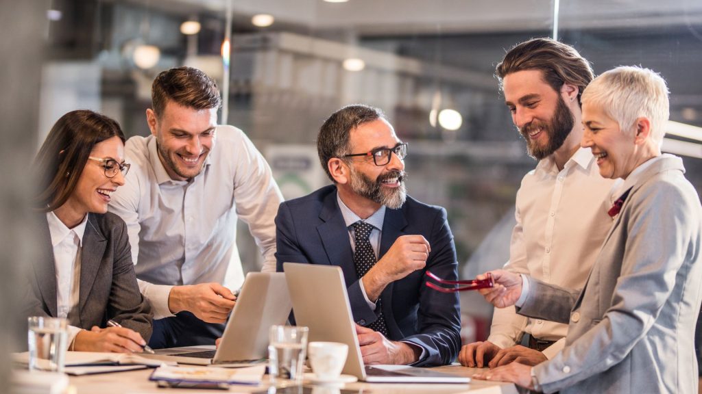 Cinco profesionales de los negocios, tres hombres y dos mujeres, sonríen y hablan alrededor de una mesa con computadores portátiles y documentos en una oficina moderna y luminosa