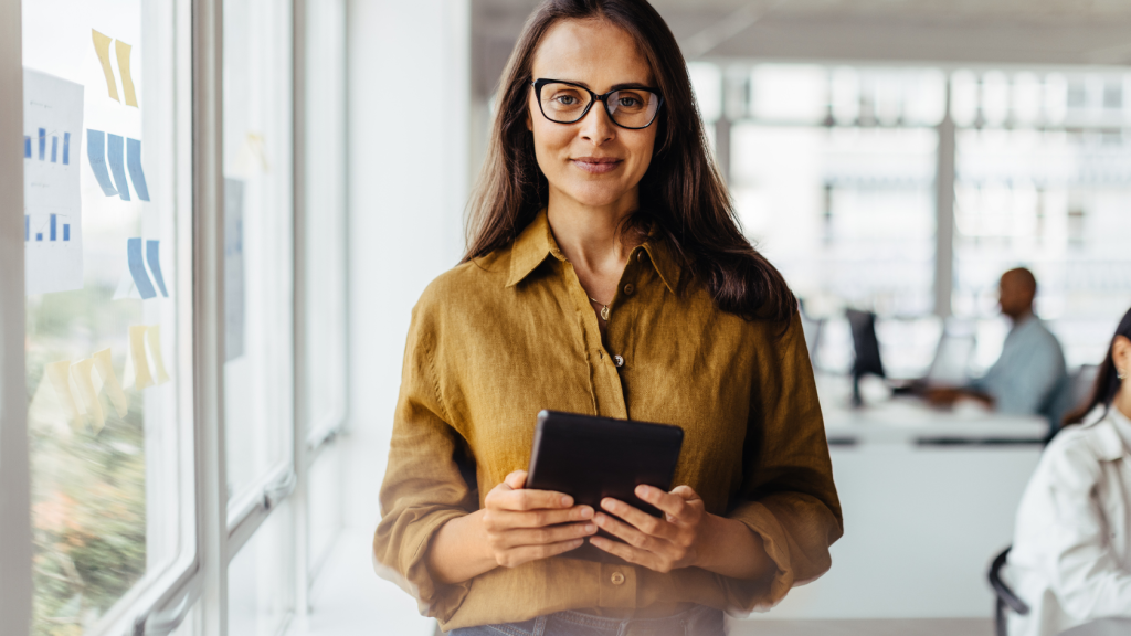 Persona de pie en una oficina luminosa, sosteniendo una tableta con ambas manos. Lleva una camisa marrón claro y el cabello largo y suelto. Al fondo, se observan colegas trabajando en escritorios y gráficos pegados en la pared izquierda